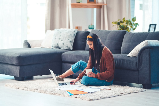 young woman on living room floor looking at laptop and papers